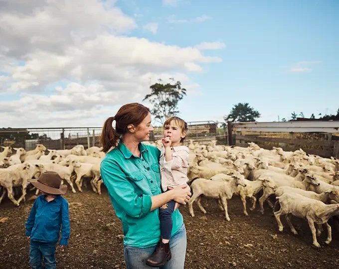 Image of a farmer and mother with her two small children, standing in a holding pen with sheep in the background.